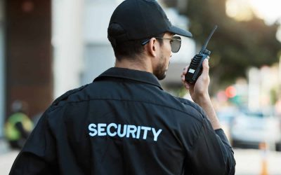 A security guard wearing a black uniform and cap, labeled "SECURITY" on the back, speaks into a walkie-talkie while on duty outdoors. The background is blurred with some people and buildings visible in the distance.