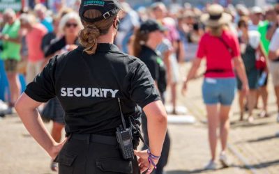 A Mobile Patrol unarmed security officer, seen from behind, wearing a black uniform with "SECURITY" written on the back, stands watch at an outdoor event in Harris County. The officer has a radio attached to their belt. A crowd of people, some walking and some standing, is gathered in the background.