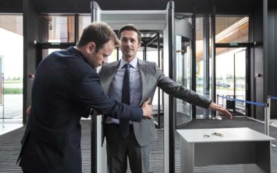 A security guard in a suit is conducting a thorough pat-down search on a man in a grey suit at a security checkpoint. The man being searched has his arms stretched out while standing in front of a metal detector, with a table nearby holding keys and a phone.