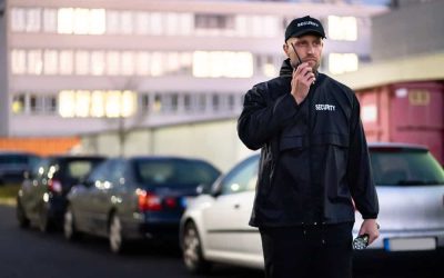 A security guard wearing a black uniform and cap labeled "Security" speaks into a two-way radio while holding a flashlight. He stands outdoors in a parking lot with several cars lined up and a building in the background during dusk or evening, providing vigilant armed security guard services for Harris County.