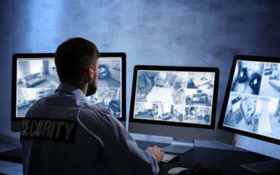 A security guard, wearing a shirt labeled "SECURITY," monitors multiple surveillance cameras displayed on three large computer screens in a dimly lit room. The screens show various locations captured by the Security Video Surveillance Harris County system.