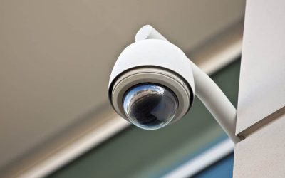 A modern, white dome security camera is mounted on the exterior corner of a building. The camera's reflective surface shows a partial view of the surrounding area, ensuring vigilant oversight alongside Mobile Patrol Harris County services. The background features a beige wall and part of a window.